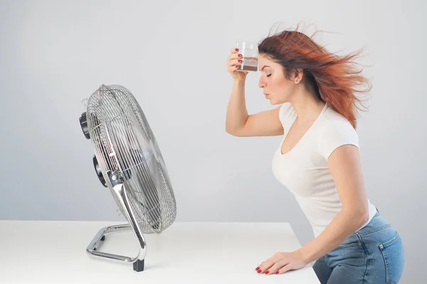 Una mujer caucásica pelirroja se enfría junto al ventilador eléctrico y bebe una bebida fría. Climatización en el apartamento — Foto de Stock