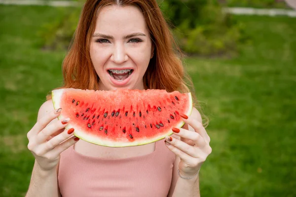 Närbild porträtt av rödhårig ung kvinna med hängslen äter vattenmelon utomhus — Stockfoto
