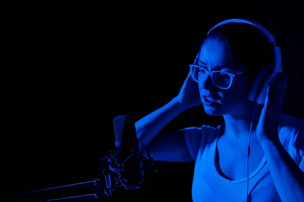 Fehér nő szemüvegben és fülhallgatóban neon fényben énekel egy mikrofonba, fekete háttérrel. Egy érzelmes lány felvesz egy dalt egy stúdióban. — Stock Fotó