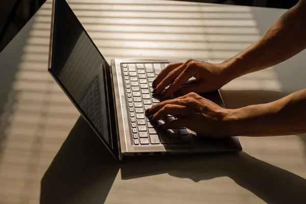 Uma mulher está digitando em um teclado de laptop em uma mesa branca. A sombra das persianas cai na área de trabalho. — Fotografia de Stock