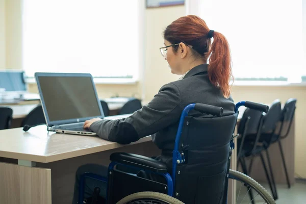 Jonge vrouw werkt op een laptop terwijl ze in een rolstoel zit in een collegezaal van de universiteit. Voorwaarden voor het onderwijzen van een gehandicapte — Stockfoto