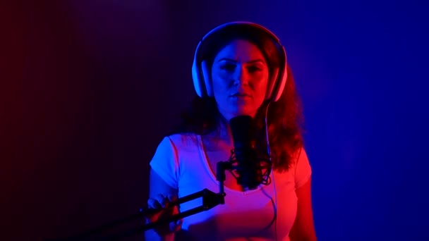 Blanke vrouw met koptelefoon zingt in een microfoon in neon licht. Een emotioneel meisje neemt een lied op in een opnamestudio. — Stockvideo