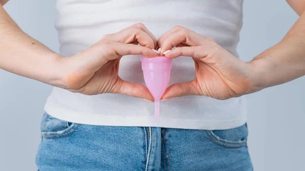 Una mujer sin rostro que usa jeans y una camiseta blanca sostiene una copa menstrual rosa sobre un fondo blanco. Manos en forma de corazón gesto — Foto de Stock