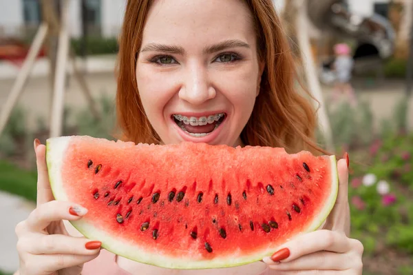Beautiful red-haired woman smiling with braces and about to eat a slice of watermelon outdoors in summer — Stock Photo, Image