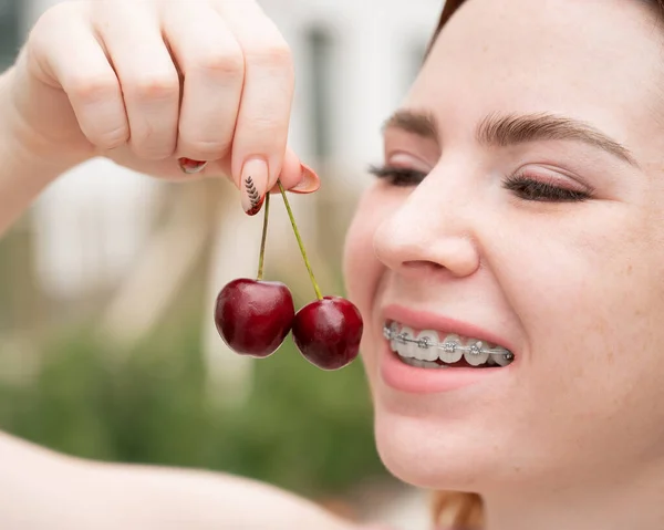 Красива молода руда жінка з брекетами на зубах їсть солодкі вишні влітку на відкритому повітрі — стокове фото