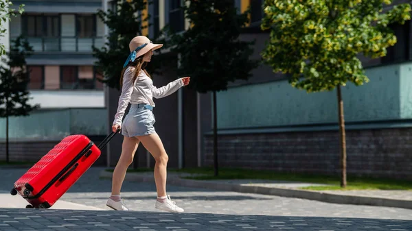 Glückliche kaukasische junge Frau mit Hut und kurzen Hosen hält einen großen roten Koffer in der Hand läuft auf einer Straße der Stadt — Stockfoto