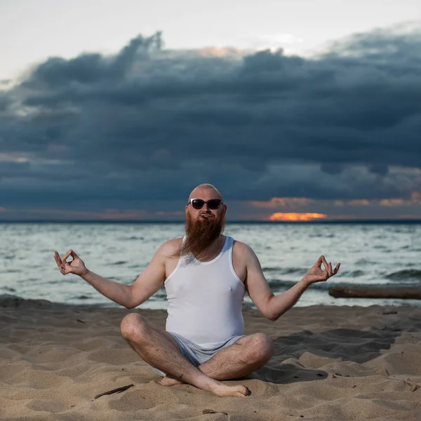 Un uomo calvo con la barba rossa pratica yoga sulla spiaggia al tramonto. Un tizio divertente con una maglietta e occhiali da sole medita sulla riva del mare. — Foto Stock
