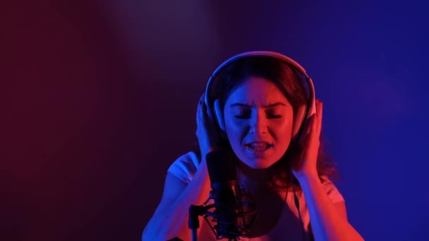 हेडफोन्समधील काकेशियन महिला निओन लाईटमध्ये मायक्रोफोनमध्ये गाते. एक भावनिक मुलगी रेकॉर्डिंग स्टुडिओमध्ये गाणे रेकॉर्ड करीत आहे — स्टॉक व्हिडिओ