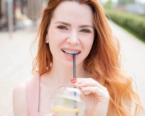Mladá krásná zrzavá žena s rovnátky pije v létě chlazenou limonádu venku. Portrét usměvavé dívky s pihami. — Stock fotografie