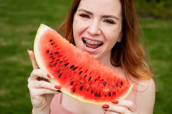 Närbild porträtt av rödhårig ung kvinna med hängslen äter vattenmelon utomhus — Stockfoto