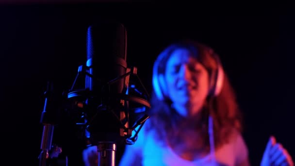 Mujer caucásica en auriculares cantando en un micrófono con luz de neón sobre un fondo negro. Una chica emocional está grabando una canción en un estudio de grabación — Vídeos de Stock