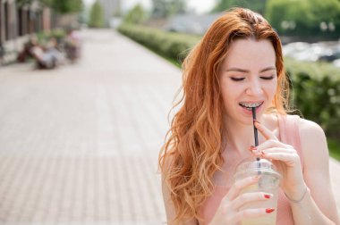 Diş teli takan güzel, kızıl saçlı bir kadın yazın dışarıda soğuk limonata içiyor. Çilli, gülümseyen bir kızın portresi..