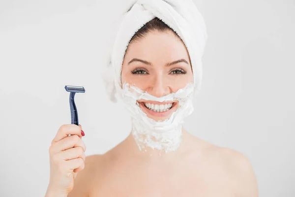 Cheerful caucasian woman with a towel on her head and shaving foam on her face holds a razor on a white background — Stock Photo, Image