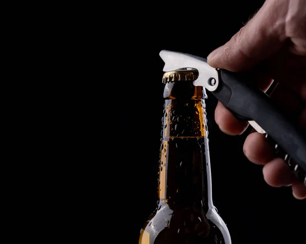 A man opens a bottle of beer from a dark glass with drops of condensation on a black background.