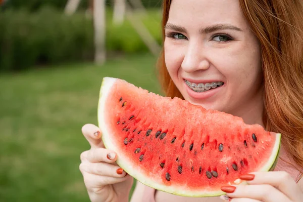 Beautiful red-haired woman smiling with braces and about to eat a slice of watermelon outdoors in summer — Stock Photo, Image