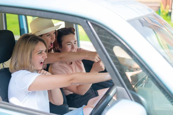 Três amigos vão numa viagem. Mulheres caucasianas dirigem em um carro e apontam os dedos de surpresa. — Fotografia de Stock