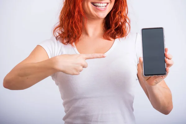 Una mujer caucásica apunta a un teléfono inteligente con una pantalla en blanco sobre un fondo blanco — Foto de Stock