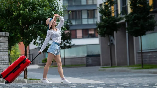 Glückliche kaukasische junge Frau mit Hut und kurzen Hosen hält einen großen roten Koffer in der Hand läuft auf einer Straße der Stadt — Stockfoto