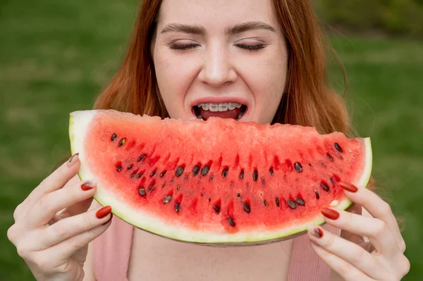 Närbild porträtt av rödhårig ung kvinna med hängslen äter vattenmelon utomhus — Stockfoto