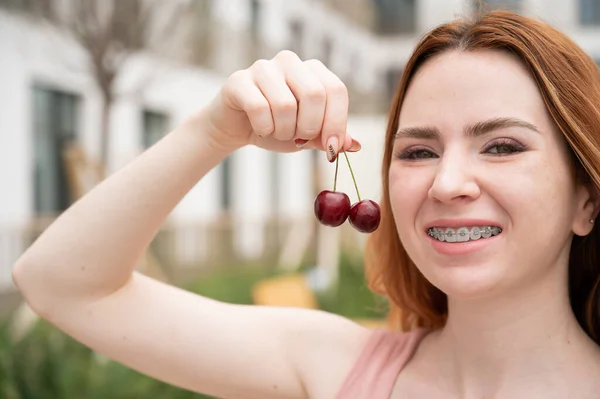 Krásná mladá zrzavá žena s rovnátky na zubech jí sladké třešně v létě venku — Stock fotografie