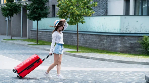 Glückliche kaukasische junge Frau mit Hut und kurzen Hosen hält einen großen roten Koffer in der Hand läuft auf einer Straße der Stadt — Stockfoto