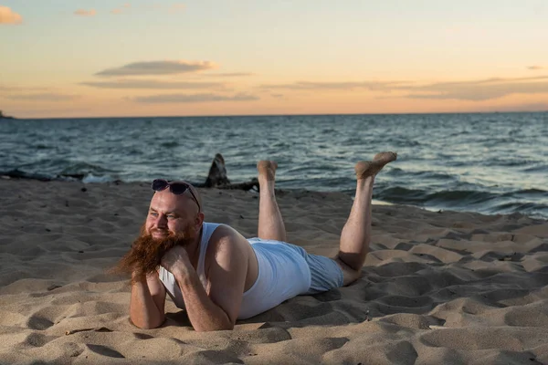 Funny bald man with red beard posing on the beach at sunset. A humorous male parody of a glamorous girl. — Stock Photo, Image