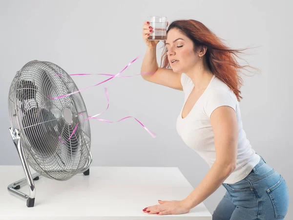 Una mujer caucásica pelirroja se enfría junto al ventilador eléctrico y bebe una bebida fría. Climatización en el apartamento — Foto de Stock