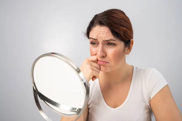 Mujer caucásica se mira en el espejo y está molesta debido a las arrugas faciales en su cara. — Foto de Stock