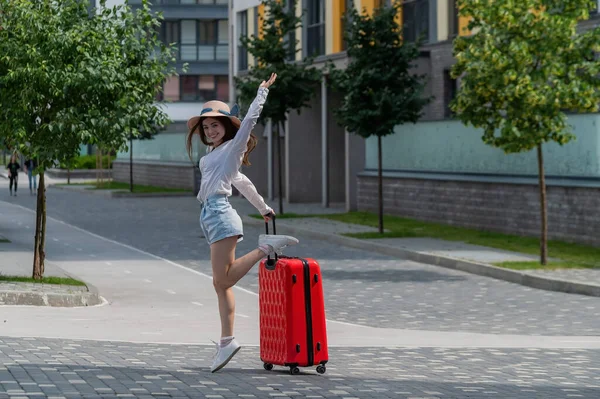 Glückliche kaukasische junge Frau mit Hut und kurzen Hosen hält einen großen roten Koffer in der Hand läuft auf einer Straße der Stadt — Stockfoto