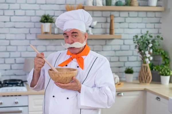 Um velho com um bigode cinzento em um chapéu de chefs está cozinhando na cozinha. — Fotografia de Stock