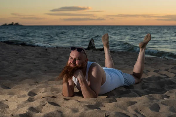 Funny bald man with red beard posing on the beach at sunset. A humorous male parody of a glamorous girl.