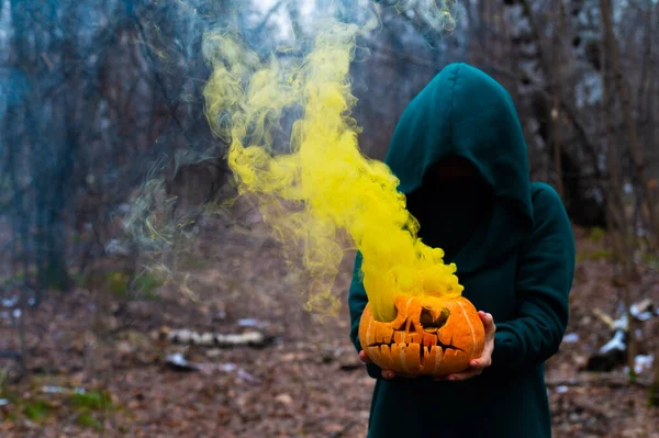 En läskig häxa håller en ångande pumpa i en djup skog. Jack o lykta avger gul rök för halloween — Stockfoto