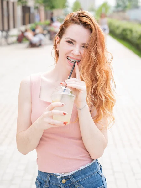 Mladá krásná zrzavá žena s rovnátky pije v létě chlazenou limonádu venku. Portrét usměvavé dívky s pihami. — Stock fotografie