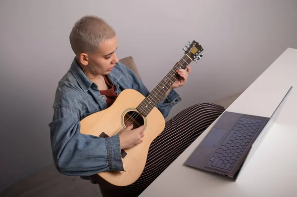 Young caucasian woman with short blonde hair playing guitar and watching training video on laptop.