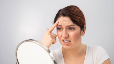 Beyaz kadın aynada kendine bakıyor ve yüzündeki kırışıklıklardan dolayı üzgün..