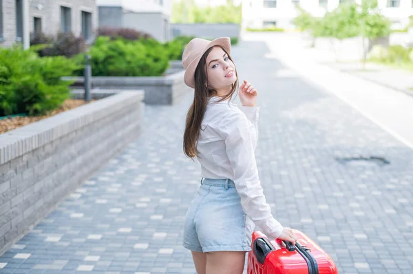 Young caucasian woman in a hat and shorts stands on the street with a red suitcase. — Stock Photo, Image