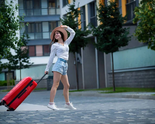 Gelukkig blanke jonge vrouw met een hoed en korte broek houdt een grote rode koffer vast loopt op een straat in de stad — Stockfoto