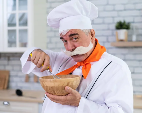 Aşçı şapkası takmış kır bıyıklı yaşlı bir adam mutfakta yemek pişiriyor.. — Stok fotoğraf