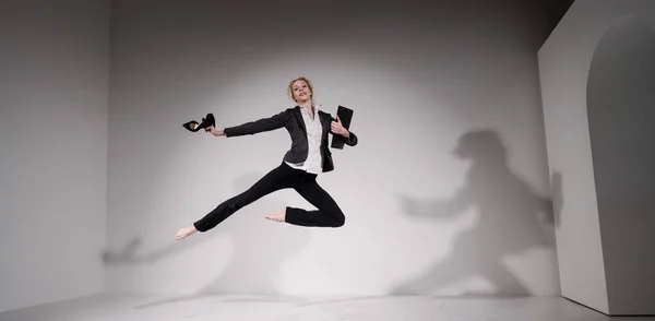 Χαριτωμένη ξυπόλητη μπαλαρίνα σε ένα επαγγελματικό κοστούμι πηδώντας με τα παπούτσια στα χέρια της σε λευκό φόντο. — Φωτογραφία Αρχείου