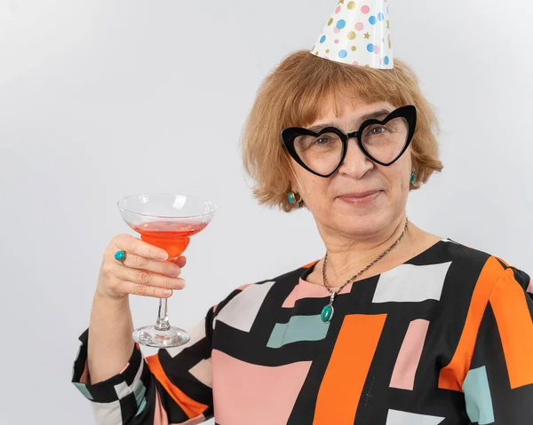 Porträtt av en leende äldre kvinna i en festlig mössa och glasögon med hjärtan dricka en cocktail på en vit bakgrund — Stockfoto