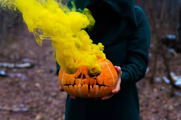 A creepy witch holds a steaming pumpkin in a deep forest. Jack o lantern emits yellow smoke for halloween — Stock Photo, Image