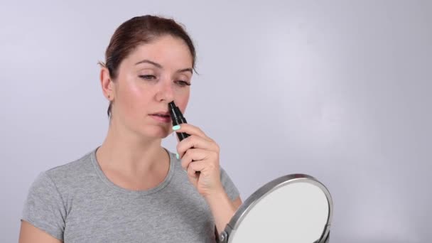 Белая женщина смотрит в зеркало и стрижет волосы в носу триммером. — стоковое видео