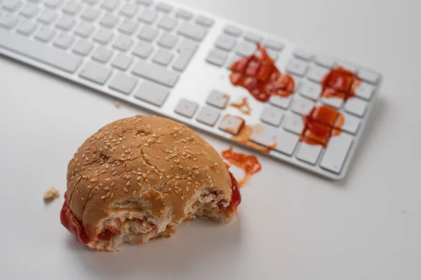 Hambúrguer e gotas de ketchup no teclado do computador na área de trabalho branca. — Fotografia de Stock