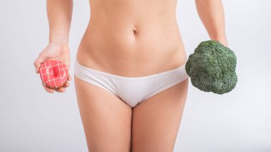 Beyaz külotlu meçhul bir kadın beyaz arka planda donut ve brokoli tutuyor. Yiyecek alışkanlıkları.