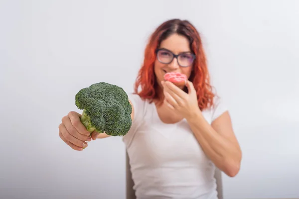 Mulher caucasiana escolhe entre legumes e fast food. Ruiva menina comer pochik e segurando brócolis em um fundo branco — Fotografia de Stock
