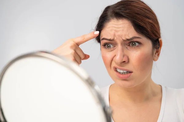 Mujer caucásica se mira en el espejo y está molesta debido a las arrugas faciales en su cara. — Foto de Stock