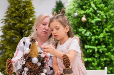 Yaşlı beyaz kadın iki torunuyla Noel için çam kozalağı süslemesi yapıyor.