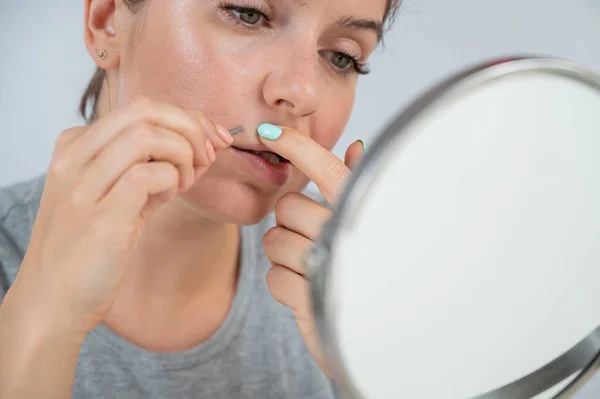 Mulher caucasiana olha no espelho e remove o bigode com a ajuda de pinças. Hirsutismo. — Fotografia de Stock