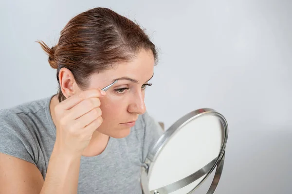 Biała kobieta patrzy w lustro i poprawia sobie brwi pęsetą — Zdjęcie stockowe