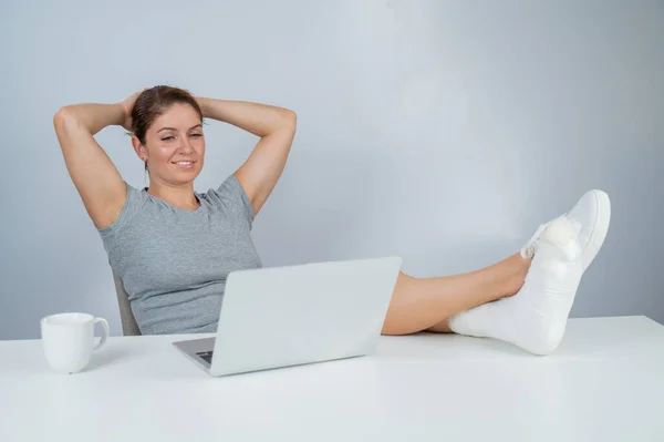 Caucasian woman lifted her leg with plaster to work desk and works on laptop on white background — Stock Photo, Image